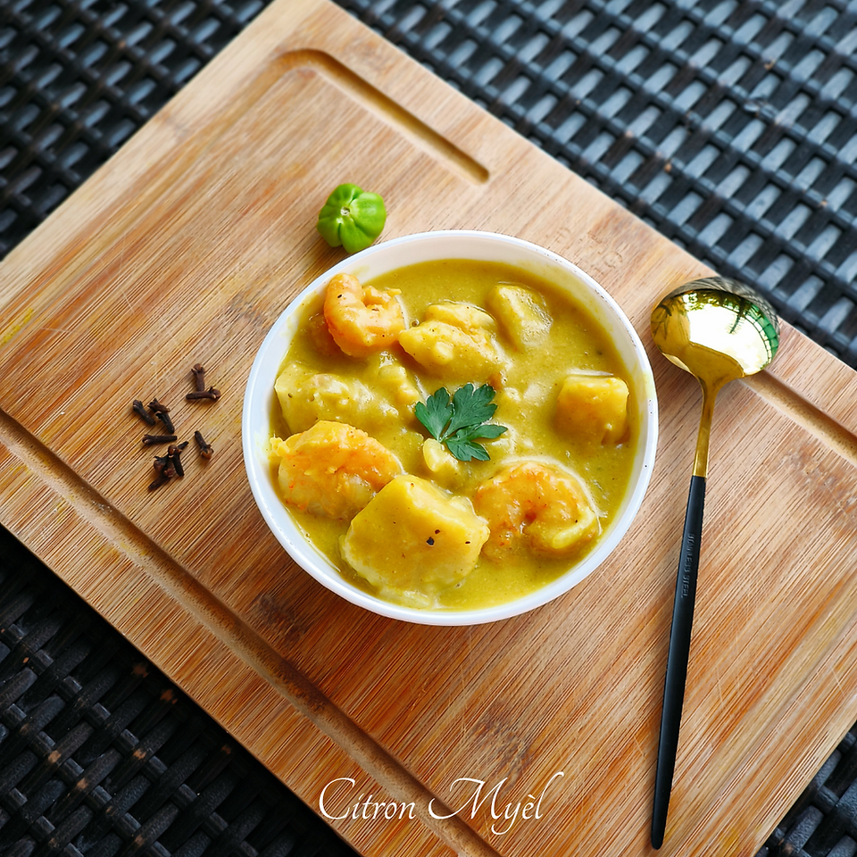 Curry de fruit à pain aux crevettes dans un bol blanc posé sur une planche en bois avecs des clous de girofle, un petit piment vert et une cuillère à soupe dorée avec le manche noir le tout sur une table tressée noire