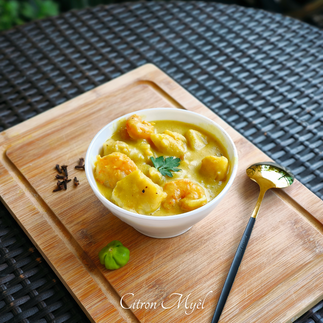 Curry de fruit à pain aux crevettes dans un bol blanc posé sur une planche en bois avecs des clous de girofle, un petit piment vert et une cuillère à soupe dorée avec le manche noir le tout sur une table tressée noire