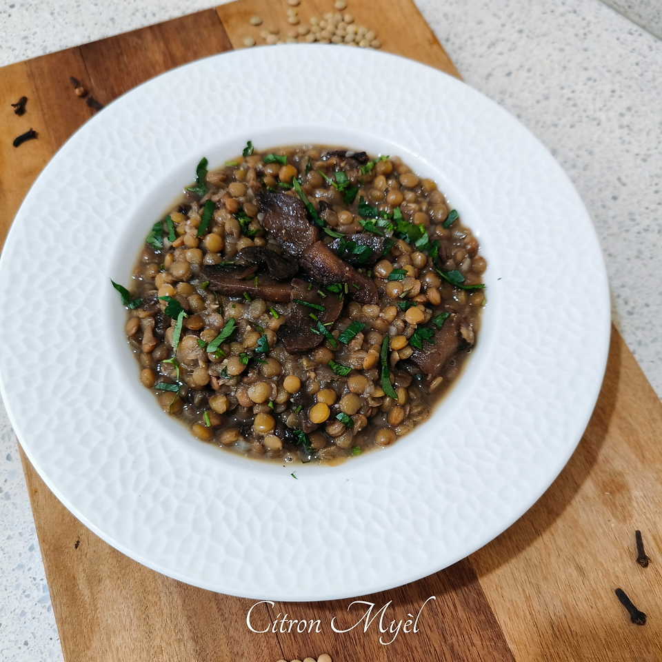 Assiette creuse blanche avec des lentilles aux champignons sur une planche en bois avec quelques tas de lentilles blondes et quelques tas de clous de girofle