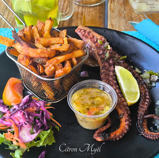 Gros plan sur une assiette noire avec un panier de frites maison, tentacules de poulpe, quartier de citorn, pot de sauce créole, et crudités (tomate, chou rouge, carotte).