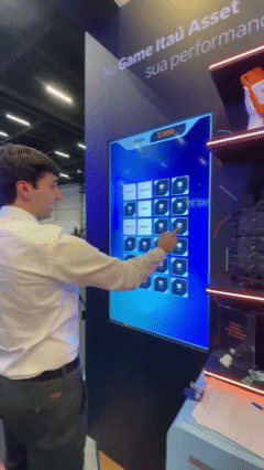 Participante interage com totem touchs Phygital Solutions em jogo de memória educativo do Itaú no XP Expert. A ativação premia de acordo com o desempenho e exibe conteúdos explicativos sobre investimentos durante a partida.