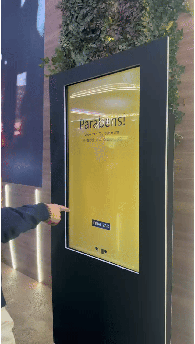 Tela vertical de 43” mostrando o game interativo “Encontre o Jeep” no Salão do Automóvel 2025. O visitante toca na tela para localizar ícones escondidos no cenário, enquanto pop-ups exibem conteúdos sobre iniciativas Jeep como Nature, Tamar e Jeep Wave.
