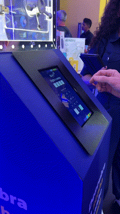 Foto de um cofre de acrílico transparente exibindo brindes, com um tablet touchscreen ao lado mostrando interface de jogo; ao fundo, um Totem Phygital com slots para mini-chaves RFID e participantes interagindo em um ambiente de convenção.