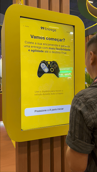 Participante joga ativação interativa da 99Entrega em um totem com joystick durante o VTEX DAY 2026, navegando em um labirinto estilo game para realizar entregas virtuais.