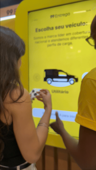 Participante joga ativação interativa da 99Entrega em um totem com joystick durante o VTEX DAY 2026, navegando em um labirinto estilo game para realizar entregas virtuais.