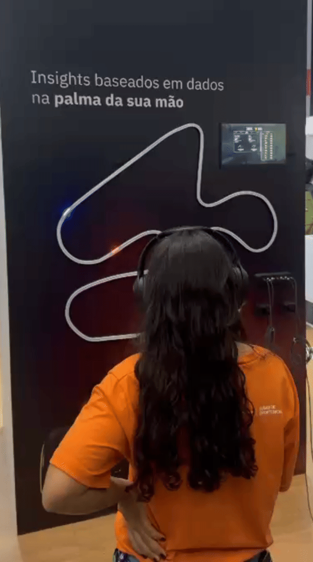 Pista de autorama personalizada da IBM com LEDs em formato de traçado, controles 3D impressos sob medida nas mãos dos participantes e estrutura técnica iluminada. Pessoas acompanham as corridas enquanto o ranking aparece em telas ao redor do estande.