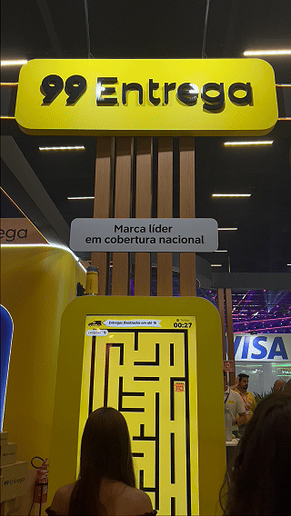 Participante joga ativação interativa da 99Entrega em um totem com joystick durante o VTEX DAY 2026, navegando em um labirinto estilo game para realizar entregas virtuais.