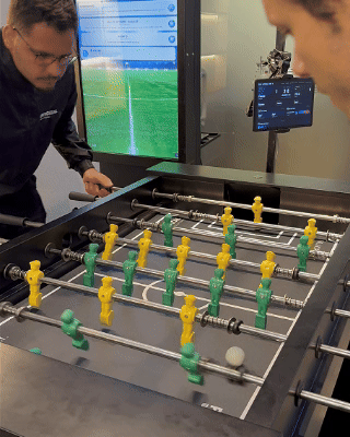 Mesa de Pebolim AI Phygital em ambiente de evento, com visual esportivo inspirado na Copa do Mundo e experiência interativa com inteligência artificial.
