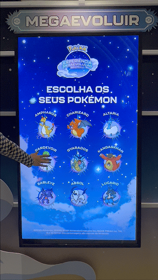 Visitantes interagem na ativação Pokémon Company | BGS 2025 – Experiência Megaevolução. Totens digitais da Phygital Solutions exibem o processo de escolha do Pokémon e da Mega Evolução, com público participando, cartões personalizados e iluminação temática do estande.