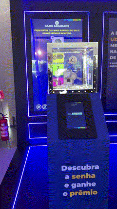 Foto de um cofre de acrílico transparente exibindo brindes, com um tablet touchscreen ao lado mostrando interface de jogo; ao fundo, um Totem Phygital com slots para mini-chaves RFID e participantes interagindo em um ambiente de convenção.