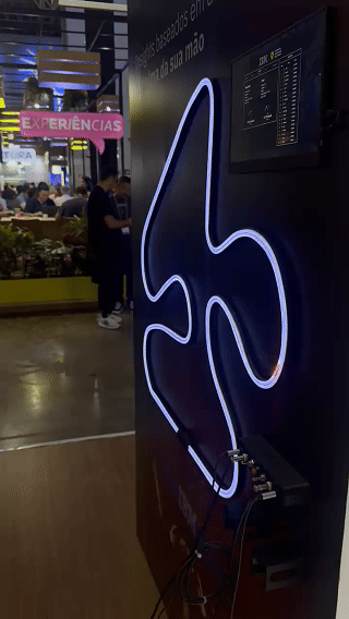 Pista de autorama personalizada da IBM com LEDs em formato de traçado, controles 3D impressos sob medida nas mãos dos participantes e estrutura técnica iluminada. Pessoas acompanham as corridas enquanto o ranking aparece em telas ao redor do estande.