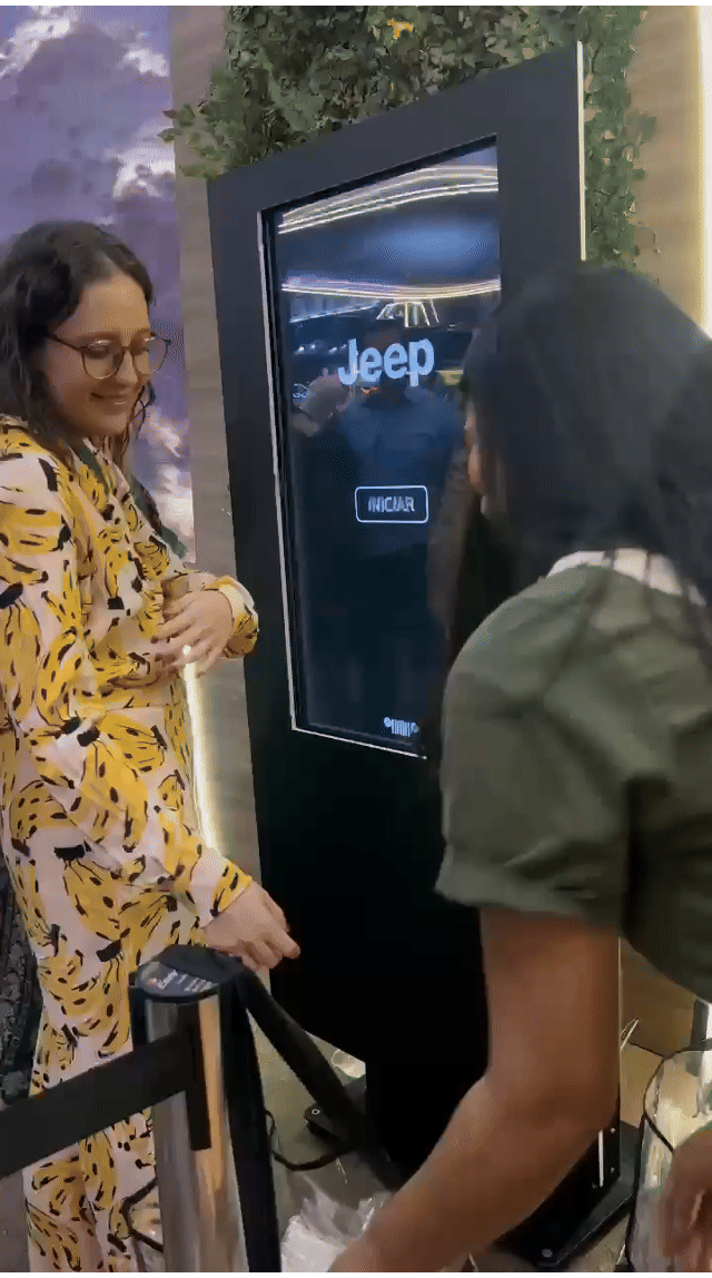 Tela vertical de 43” mostrando o game interativo “Encontre o Jeep” no Salão do Automóvel 2025. O visitante toca na tela para localizar ícones escondidos no cenário, enquanto pop-ups exibem conteúdos sobre iniciativas Jeep como Nature, Tamar e Jeep Wave.