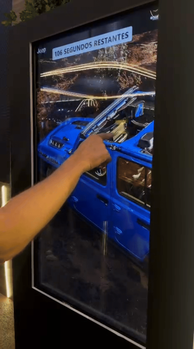 Tela vertical de 43” mostrando o game interativo “Encontre o Jeep” no Salão do Automóvel 2025. O visitante toca na tela para localizar ícones escondidos no cenário, enquanto pop-ups exibem conteúdos sobre iniciativas Jeep como Nature, Tamar e Jeep Wave.