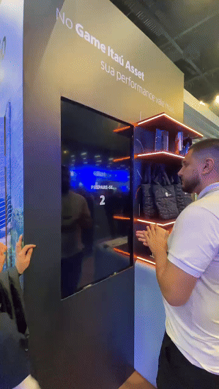 Participante interage com totem touchs Phygital Solutions em jogo de memória educativo do Itaú no XP Expert. A ativação premia de acordo com o desempenho e exibe conteúdos explicativos sobre investimentos durante a partida.