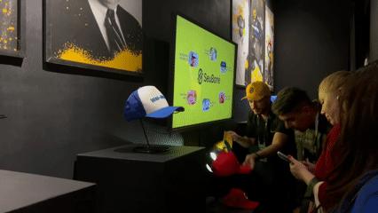 Visitantes interagem com o Museu RFID do Seu Boné na Brazil Promotion 2025, posicionando bonés históricos com tags RFID sobre suporte personalizado produzido em impressoras 3D pela Phygital Solutions, revelando histórias e curiosidades em tempo real.