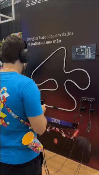 Pista de autorama personalizada da IBM com LEDs em formato de traçado, controles 3D impressos sob medida nas mãos dos participantes e estrutura técnica iluminada. Pessoas acompanham as corridas enquanto o ranking aparece em telas ao redor do estande.