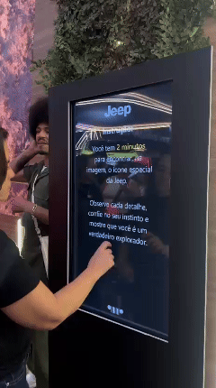 Tela vertical de 43” mostrando o game interativo “Encontre o Jeep” no Salão do Automóvel 2025. O visitante toca na tela para localizar ícones escondidos no cenário, enquanto pop-ups exibem conteúdos sobre iniciativas Jeep como Nature, Tamar e Jeep Wave.