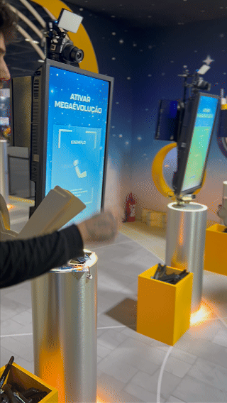 Visitantes interagem na ativação Pokémon Company | BGS 2025 – Experiência Megaevolução. Totens digitais da Phygital Solutions exibem o processo de escolha do Pokémon e da Mega Evolução, com público participando, cartões personalizados e iluminação temática do estande.