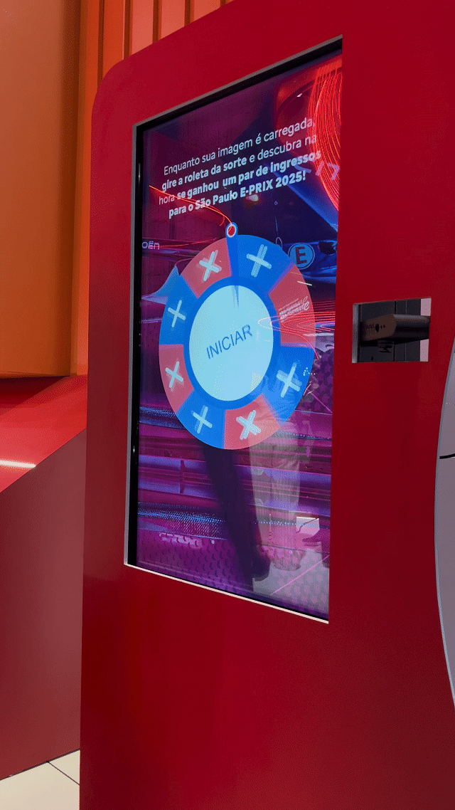 Visitante sendo fotografado em um totem da Citroën no Salão do Automóvel 2025, tendo sua imagem transformada por IA em um piloto da Fórmula E. A foto final aparece na tela ao lado, enquanto outro participante interage com a Roleta Digital personalizada com identidade da marca.