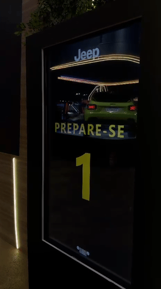 Tela vertical de 43” mostrando o game interativo “Encontre o Jeep” no Salão do Automóvel 2025. O visitante toca na tela para localizar ícones escondidos no cenário, enquanto pop-ups exibem conteúdos sobre iniciativas Jeep como Nature, Tamar e Jeep Wave.