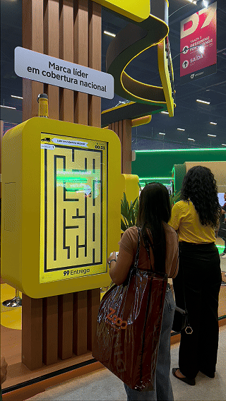 Participante joga ativação interativa da 99Entrega em um totem com joystick durante o VTEX DAY 2026, navegando em um labirinto estilo game para realizar entregas virtuais.