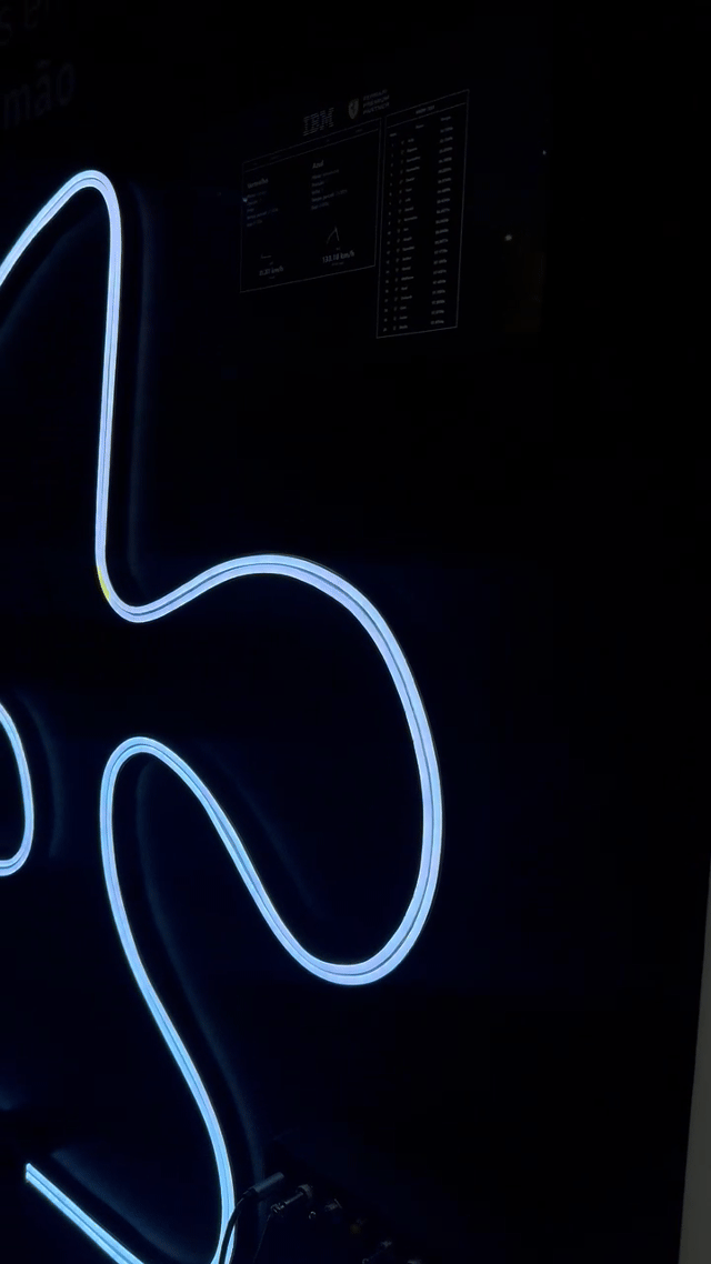 Pista de autorama personalizada da IBM com LEDs em formato de traçado, controles 3D impressos sob medida nas mãos dos participantes e estrutura técnica iluminada. Pessoas acompanham as corridas enquanto o ranking aparece em telas ao redor do estande.