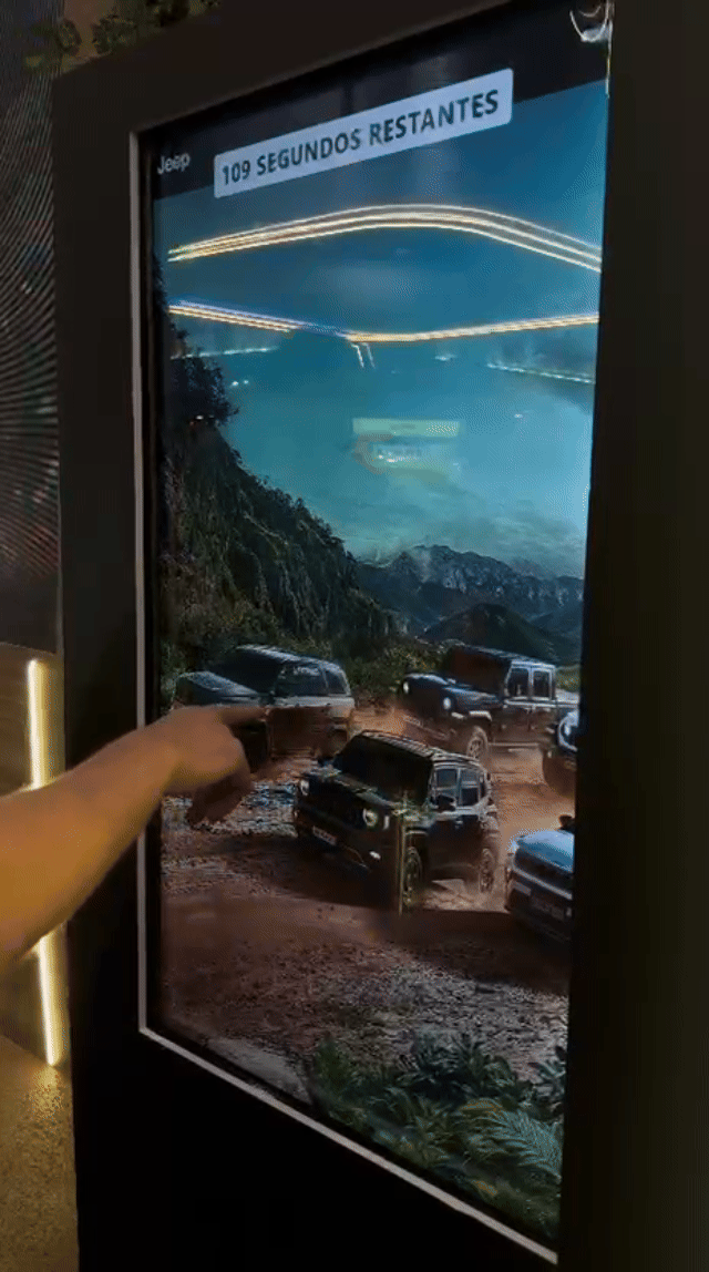 Tela vertical de 43” mostrando o game interativo “Encontre o Jeep” no Salão do Automóvel 2025. O visitante toca na tela para localizar ícones escondidos no cenário, enquanto pop-ups exibem conteúdos sobre iniciativas Jeep como Nature, Tamar e Jeep Wave.