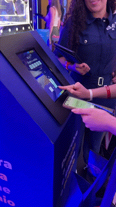 Foto de um cofre de acrílico transparente exibindo brindes, com um tablet touchscreen ao lado mostrando interface de jogo; ao fundo, um Totem Phygital com slots para mini-chaves RFID e participantes interagindo em um ambiente de convenção.