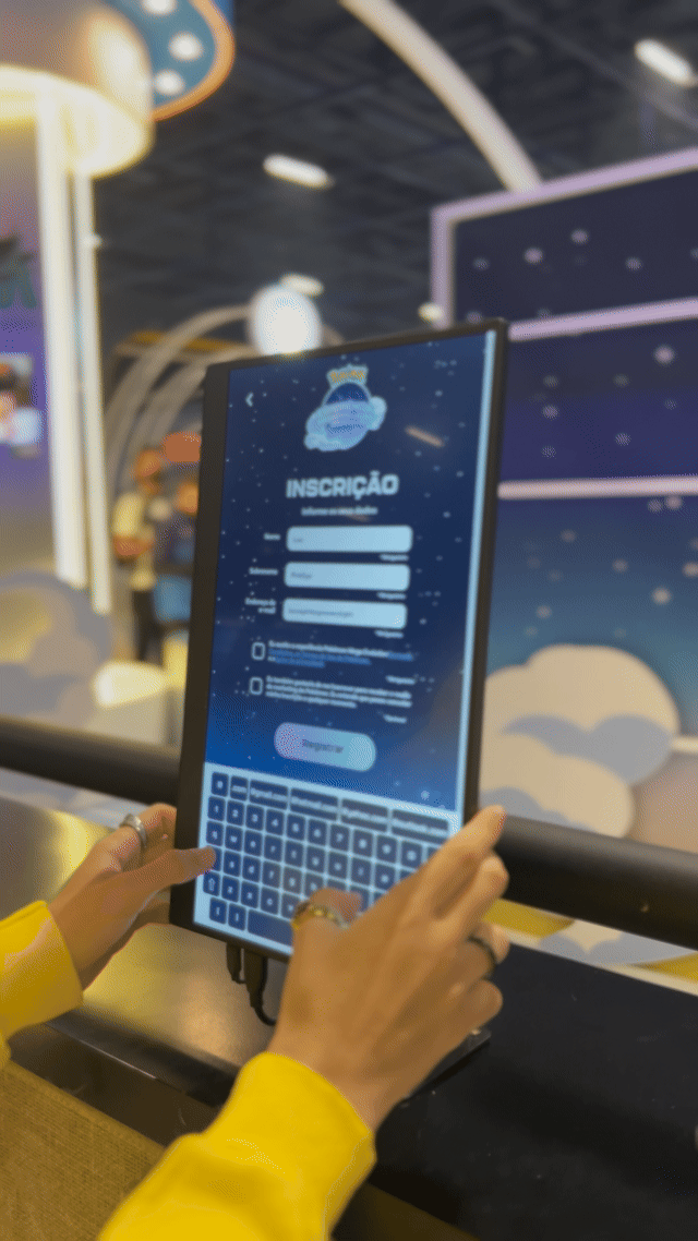 Visitantes interagem na ativação Pokémon Company | BGS 2025 – Experiência Megaevolução. Totens digitais da Phygital Solutions exibem o processo de escolha do Pokémon e da Mega Evolução, com público participando, cartões personalizados e iluminação temática do estande.
