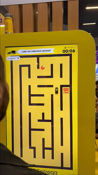 Participante joga ativação interativa da 99Entrega em um totem com joystick durante o VTEX DAY 2026, navegando em um labirinto estilo game para realizar entregas virtuais.