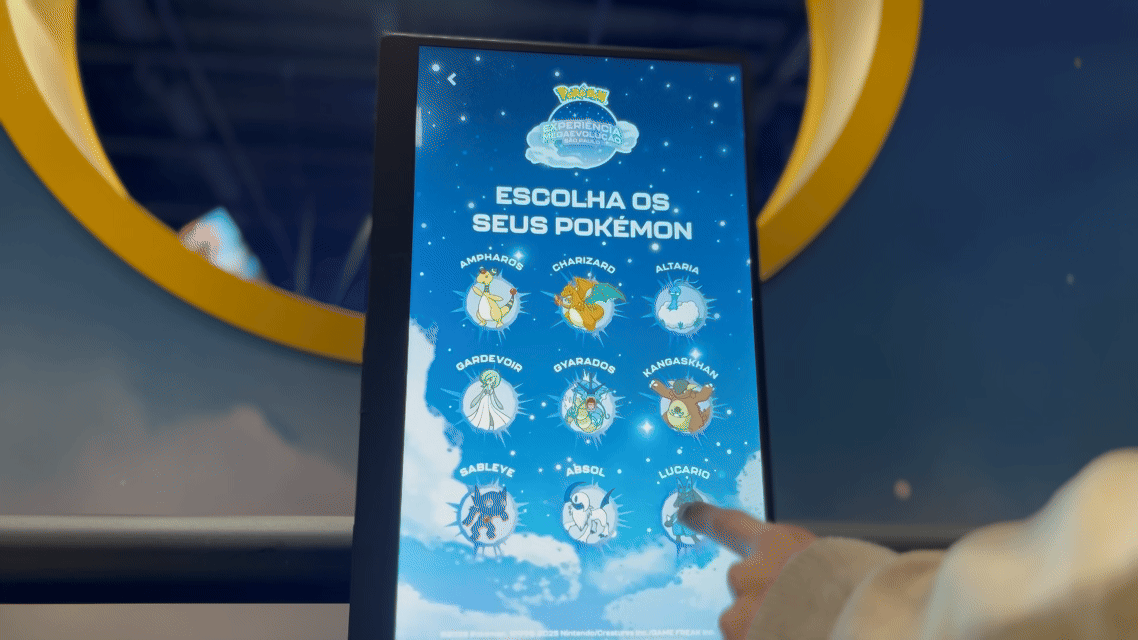 Visitantes interagem na ativação Pokémon Company | BGS 2025 – Experiência Megaevolução. Totens digitais da Phygital Solutions exibem o processo de escolha do Pokémon e da Mega Evolução, com público participando, cartões personalizados e iluminação temática do estande.