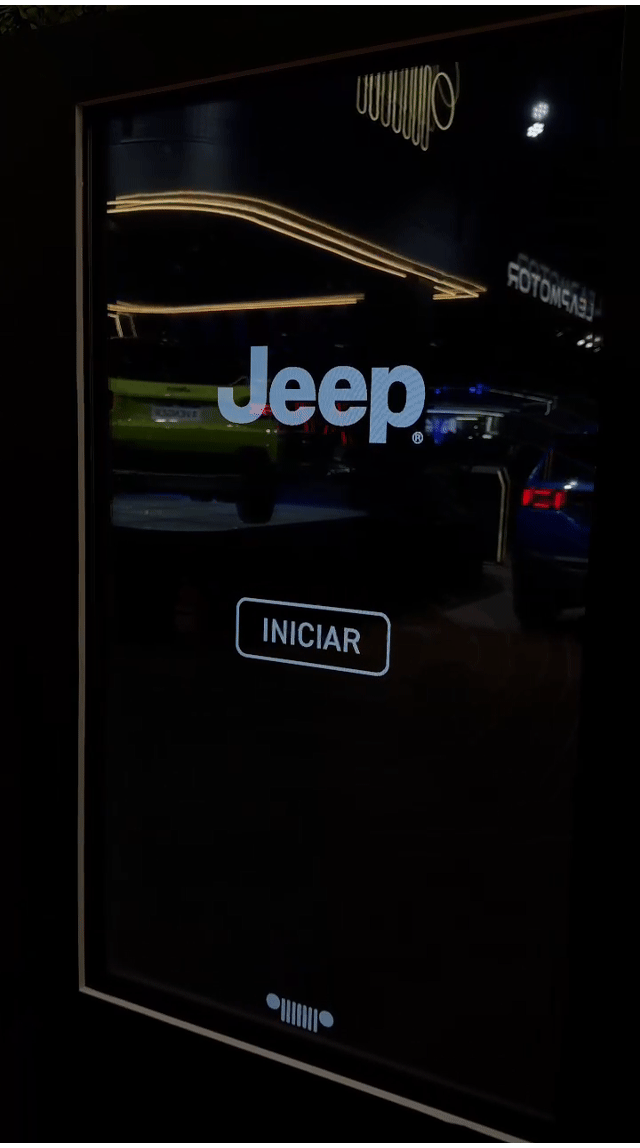 Tela vertical de 43” mostrando o game interativo “Encontre o Jeep” no Salão do Automóvel 2025. O visitante toca na tela para localizar ícones escondidos no cenário, enquanto pop-ups exibem conteúdos sobre iniciativas Jeep como Nature, Tamar e Jeep Wave.