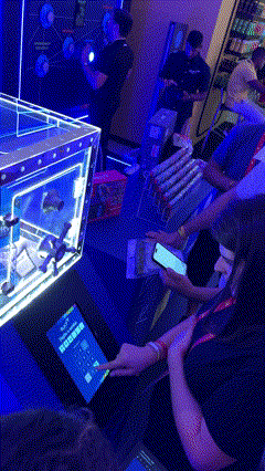 Foto de um cofre de acrílico transparente exibindo brindes, com um tablet touchscreen ao lado mostrando interface de jogo; ao fundo, um Totem Phygital com slots para mini-chaves RFID e participantes interagindo em um ambiente de convenção.