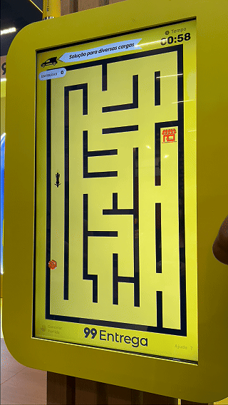 Participante joga ativação interativa da 99Entrega em um totem com joystick durante o VTEX DAY 2026, navegando em um labirinto estilo game para realizar entregas virtuais.