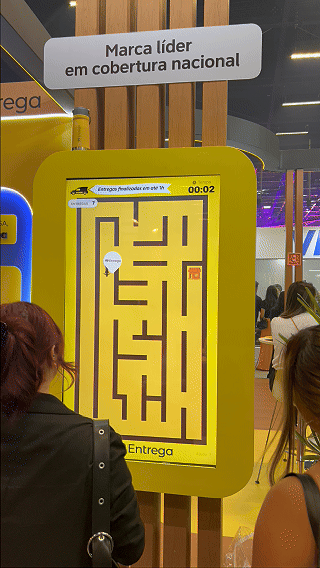 Participante joga ativação interativa da 99Entrega em um totem com joystick durante o VTEX DAY 2026, navegando em um labirinto estilo game para realizar entregas virtuais.