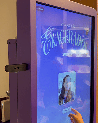 Pessoa interagindo com totem digital em loja da Vivo, realizando cadastro e tirando foto para geração de card personalizado impresso na hora durante ação pré-Lollapalooza.