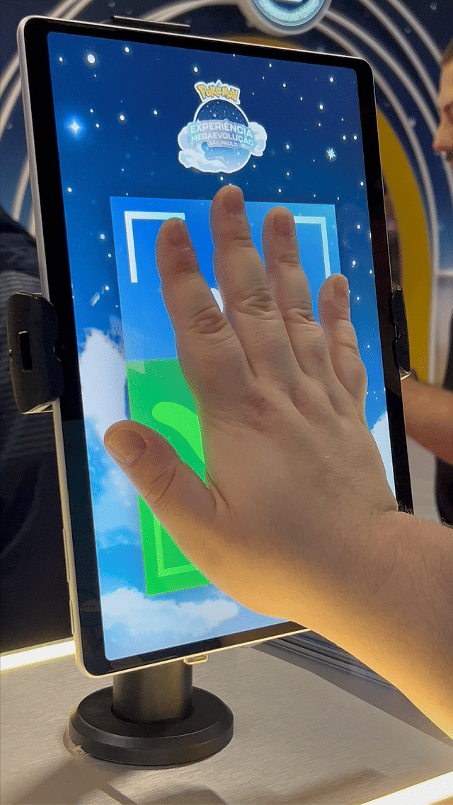Visitantes interagem na ativação Pokémon Company | BGS 2025 – Experiência Megaevolução. Totens digitais da Phygital Solutions exibem o processo de escolha do Pokémon e da Mega Evolução, com público participando, cartões personalizados e iluminação temática do estande.
