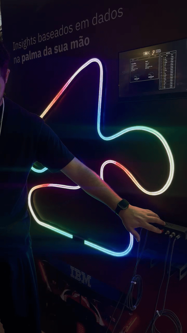 Pista de autorama personalizada da IBM com LEDs em formato de traçado, controles 3D impressos sob medida nas mãos dos participantes e estrutura técnica iluminada. Pessoas acompanham as corridas enquanto o ranking aparece em telas ao redor do estande.