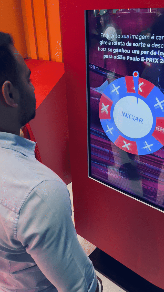 Visitante sendo fotografado em um totem da Citroën no Salão do Automóvel 2025, tendo sua imagem transformada por IA em um piloto da Fórmula E. A foto final aparece na tela ao lado, enquanto outro participante interage com a Roleta Digital personalizada com identidade da marca.