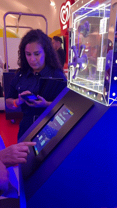 Foto de um cofre de acrílico transparente exibindo brindes, com um tablet touchscreen ao lado mostrando interface de jogo; ao fundo, um Totem Phygital com slots para mini-chaves RFID e participantes interagindo em um ambiente de convenção.
