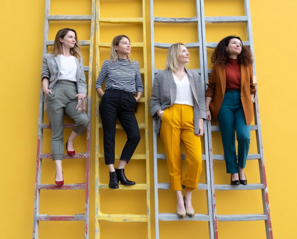 Four women in stylish outfits stand on ladders against a vibrant yellow wall, conveying confidence and camaraderie.