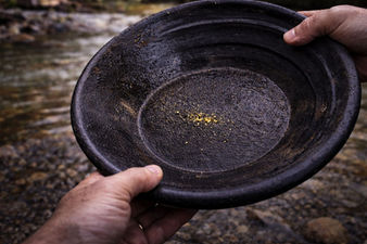 Close-up of hands holding a wet black gold pan beside a rocky stream, with a small amount of fine gold mixed into dark magnetite (“black sand”) in the bottom.