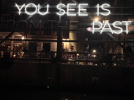 Enseigne lumineuse au néon indiquant "All you see is Past", reflectée sur l'eau sombre la nuit, avec des bâtiments doucement éclairés en arrière plan, au centre d'Amsterdam.