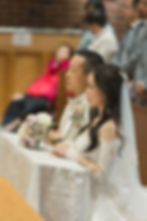 Bride and groom sit at a decorated altar, holding flowers, in a church ceremony. Attendees in the background. The mood is solemn/St. John the Apostle Parish wedding ceremony Vancouver