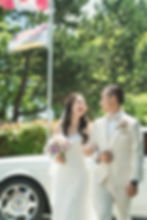 Bride and groom in white attire walking arm-in-arm, smiling. She holds a bouquet. Flags and greenery in the sunny background. Romantic mood /Quilchena Golf Club wedding photos Richmond