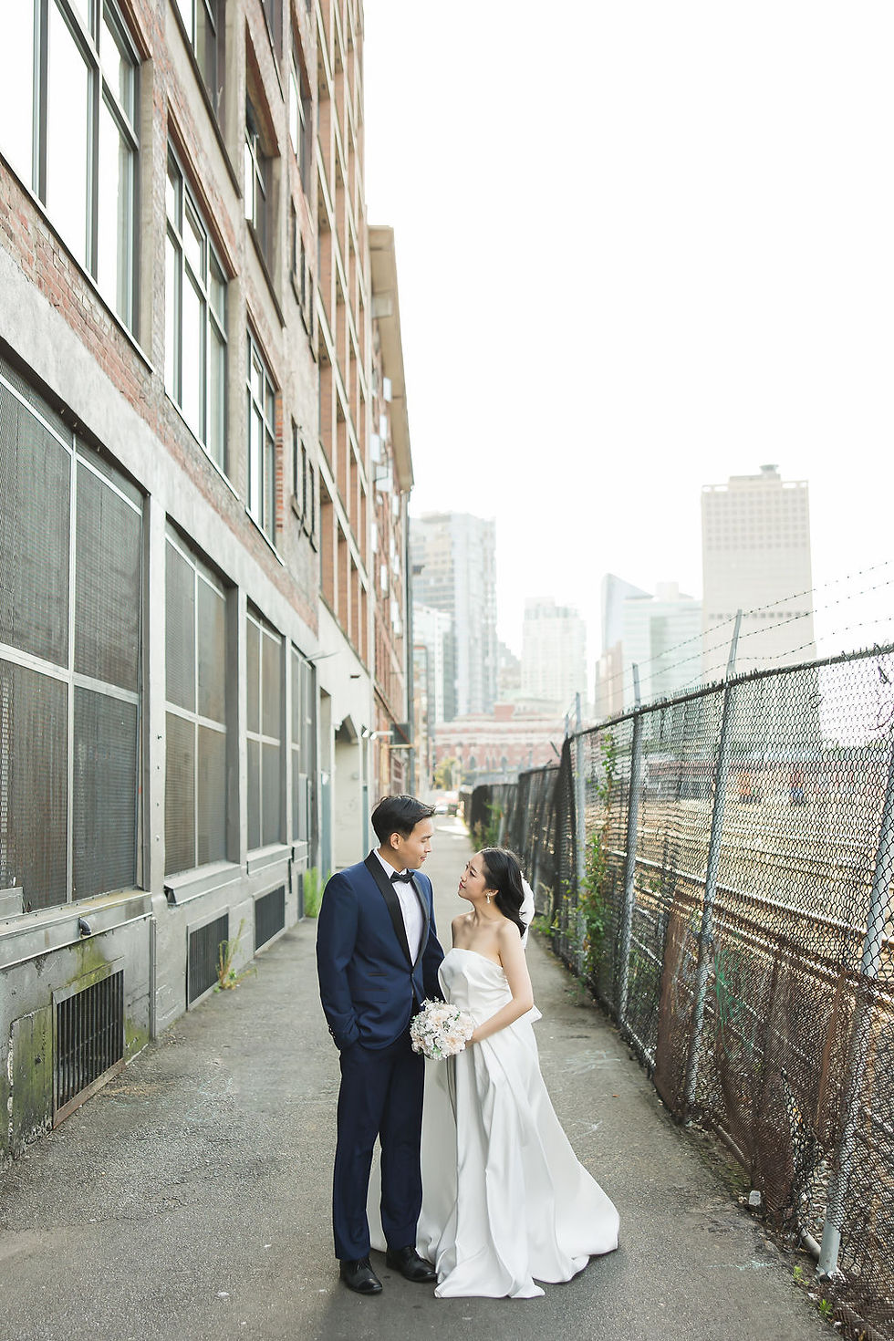 Bride and groom stand on an urban street, bride in white gown holding flowers, groom in navy suit, with city buildings in background. Pre Wedding Photoshoot Vancouver – One-Stop Gastown Experience with The Eunice & Co.