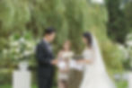 Bride and groom exchange vows outdoors, surrounded by white flowers. An officiant smiles behind them. Lush greenery sets a serene mood at VanDusen Botanical Garden Wedding and Shaughnessy Restaurant wedding