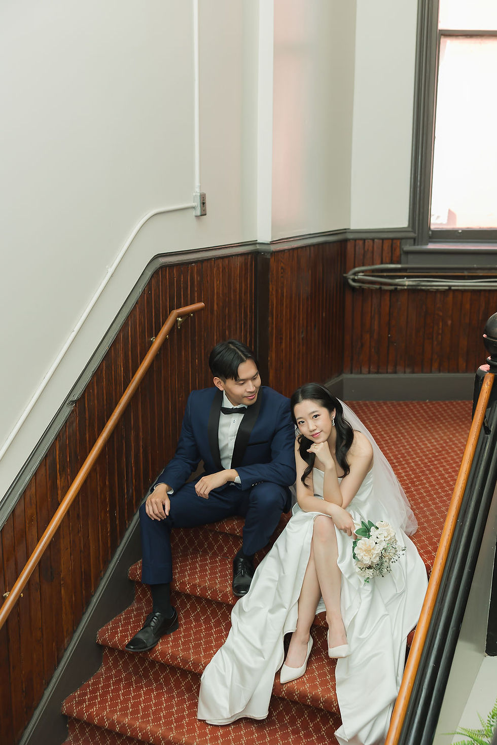 Bride and groom sit on red-carpeted stairs; she wears a white dress holding flowers, he’s in a blue suit. Wooden-paneled walls in background. Pre Wedding Photoshoot Vancouver – One-Stop Gastown Experience with The Eunice & Co.