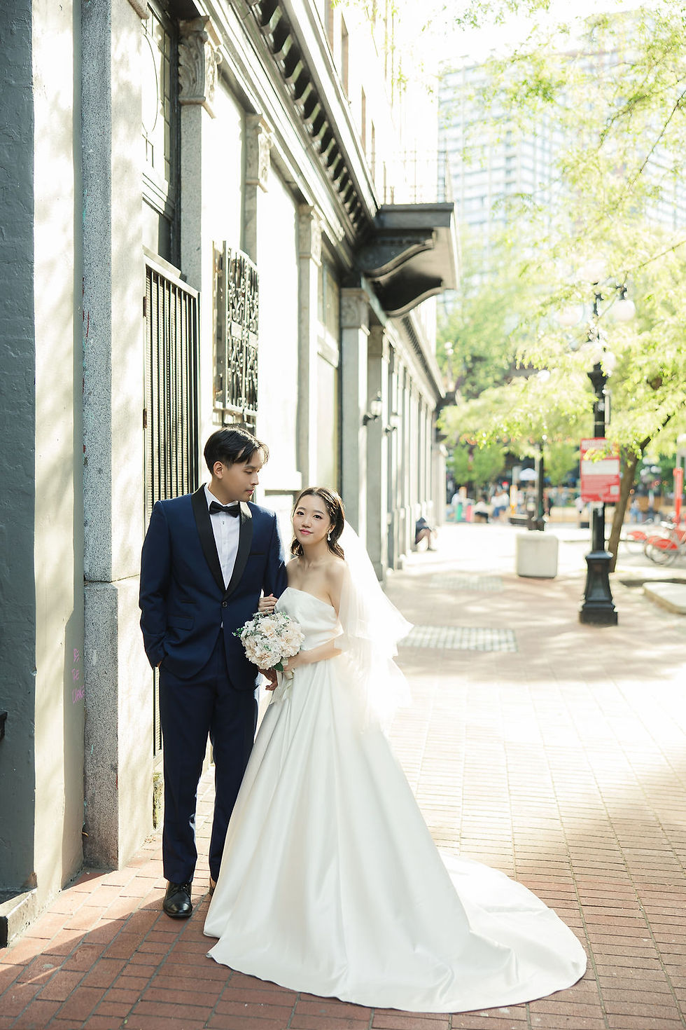 Bride in white gown and groom in blue suit stand arm in arm on a sunlit street with trees and buildings. She holds a bouquet. Pre Wedding Photoshoot Vancouver – One-Stop Gastown Experience with The Eunice & Co.