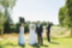 Bride and groom stand with groomsman and bridesmaid in a sunny garden. Bride holds bouquet; greenery and a small river in the background /Mayfair Golf Course Wedding